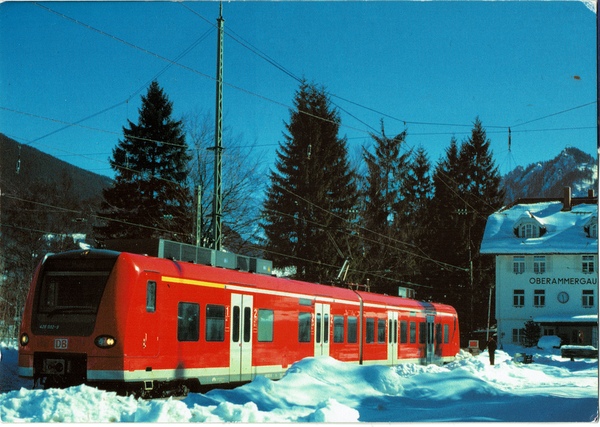 Man sieht einen rot-weiße Elektrotriebwagen der DB Regio an einem Bahnhof stehen vor blauem Himmel. Es ist Winter und man kann kleinere Schneehaufen und Schneeverwehungen sehen. Im Hintergrund sieht man die Berge, wie auch das Bahnhofsgebäude von Oberammergau. Die Uhr am Gebäude zeigt halb zwölf.