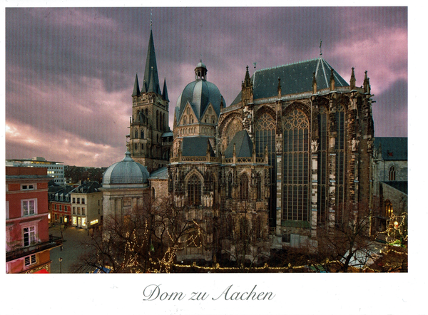 In einer abendlichen, festlichen Stimmung ist der Aachener Dom zu sehen. Er steht in vielen bräunlichen, gräulichen Schattierungen vor einem rot-violett-grauen Himmel. In der Umgebung sind einige Geschäfte und andere Häuser zu sehen. Einige Bäume. Vereinzelt sieht man erleuchtete Fenster und in den Bäumen Lichterketten.