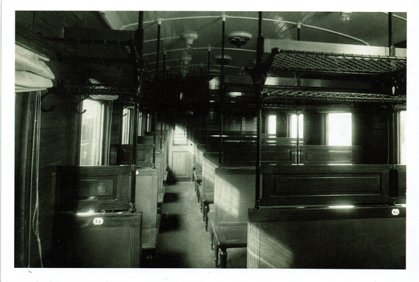 Man sieht ein schwarz-weiß Foto eines alten Eisenbahnwagenabteil mit Holzsitze. Auch wenn die Bänke sehr hart aussehen, wirkt es gemütlich.