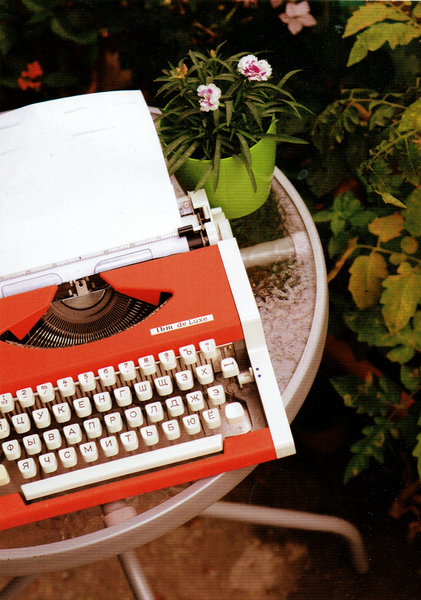 Eine rot-weiße Schreibmaschine mit eingespanntem Papier steht auf einem Tisch in mitten von Blumen.