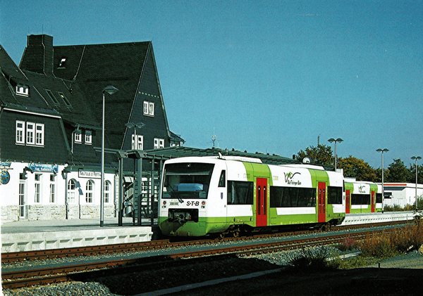 Man sieht einen Triebwagen der STB in grün-weiß mit roten Türen vor dem Bahnhofsgebäude von Neuhaus stehen. Das Gebäude ist frisch saniert und hat weiße Wände, sowie eine schöne Schiefer Eindeckung. Es ist sonnig und die Himmel blau