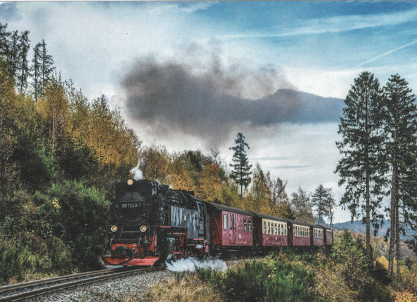 Eine schwarze Dampflok mit mäßigen Rauch zieht einie Wagen in braun-beige der Harzer Schmalspurbahnen einen kleinen Hügel (vermutlich den Broken) hoch. Man kann Bäume links und rechts von der strecke sehen. Die Karte wirklich kurz vor gezeichnet auf Grund der Druckmethode.