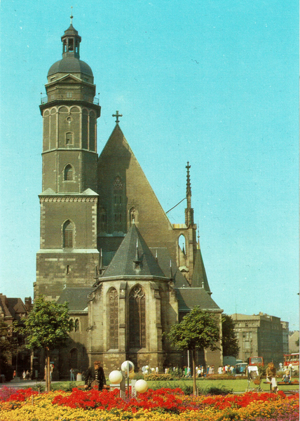 Man sihet auf eine älteren Karte, ca 1980, eine Kirche -- die Thomaskirche -- mit grau-braunen Mauerwerk, verputzt. Davor eine wieso mit roten Blumen, einer Art Lampen-Installation in Form einer Blume und natürlich Menschen. Der Himmel ist blau